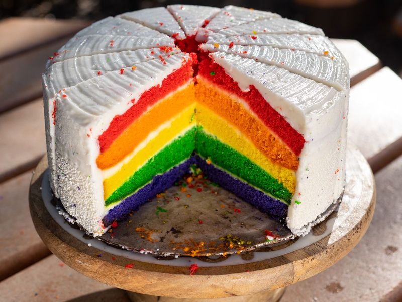 Angeschnittene Regenbogentorte mit weisser Glasur auf einem Holzständer, im Anschnitt leuchten die bunten Schichten im Sonnenlicht