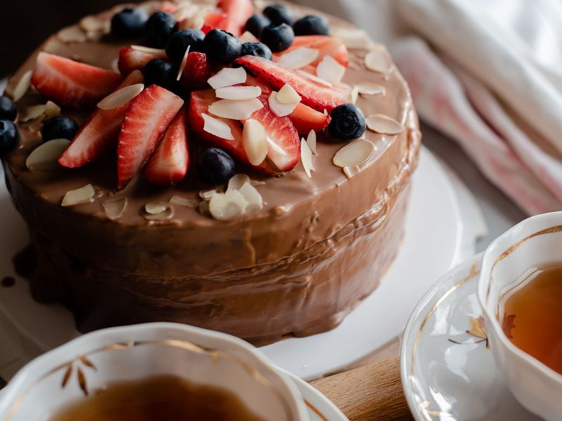 Schokoladig glasierte Torte mit frischen Erdbeeren, Heidelbeeren und Mandelblättchen, daneben zwei Tassen Tee